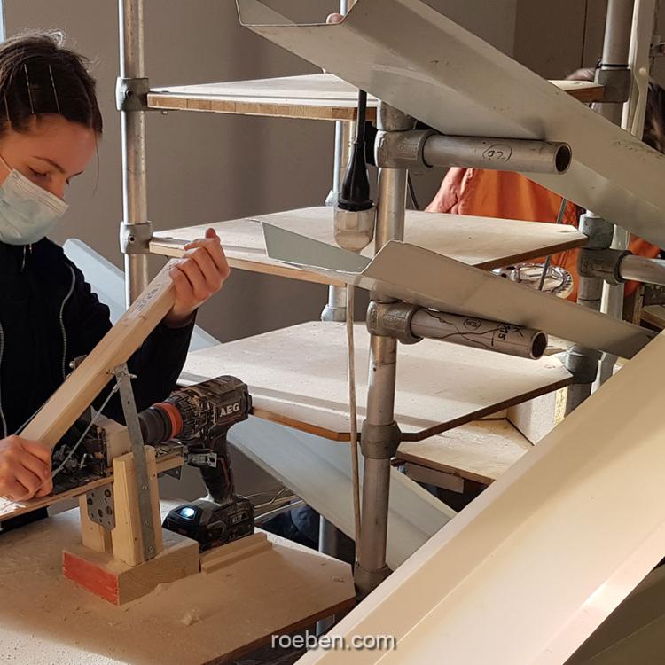 Frau mit Maske arbeitet an einer Holzstruktur in einem hellen Raum.