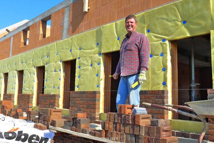 Die zweischalige Wand: Nachhaltige Zukunftskonstruktion Bauarbeiter mit Flanellhemd lächelt auf Baustelle neben Ziegelmauerwerk.