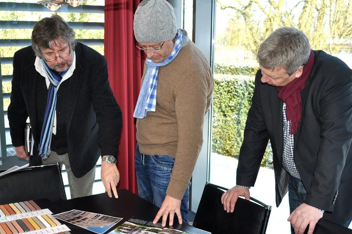 Drei Männer in Winterkleidung betrachten Materialmuster an einem Tisch.
