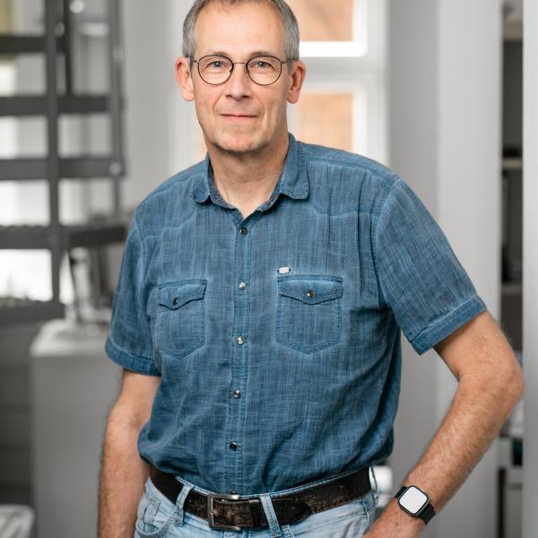 Architekt Ulrich Wolbeck mit Brille in Jeanshemd steht lächelnd in hellem Raum.