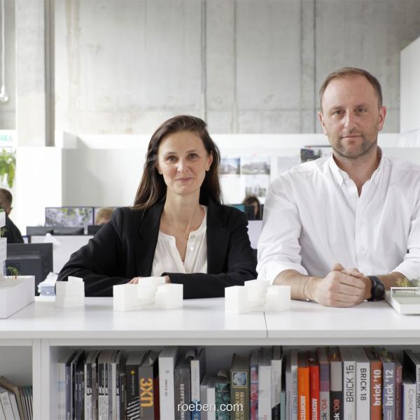 Isabel van Haute, ThomasColdefy stehen hinter einem Tisch in einem modernen Büro.