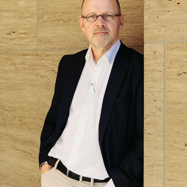 Tobias Nöfer im Anzug lehnt an beiger Steinwand, schaut ernst.