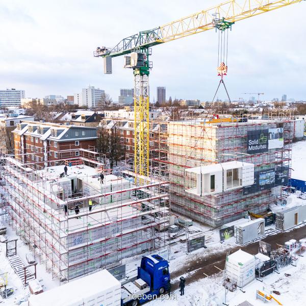 Bauarbeiten im Schnee mit Kran und Gerüst, moderne Stadt im Hintergrund.