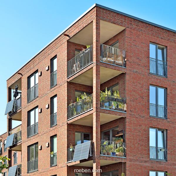 Mehrstöckiges Backsteingebäude mit Balkonen und Pflanzen, blauer Himmel.