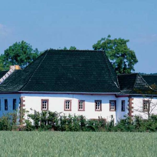 Die dunkle Farbe des Tondachziegels steht im wohltuenden Kontrast zur weißen Fassade mit ihren braun-rot abgesetzten Fenster- und Türeinfassungen. Die glänzende Oberfläche des Ziegels spielt mit dem Blau des Himmels und gibt so dem historischen G